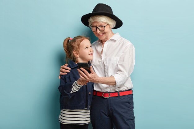 Elderly lady and small cute girl use mobile photo for viewing photos, sending messages, embrace with love each other, spend free time together. Little kid teaches grandma how use cell phone.