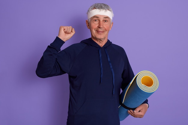 Elderly man clenching fist and holding yoga mat, wearing sport clothing and headband, posing isolated over lilac