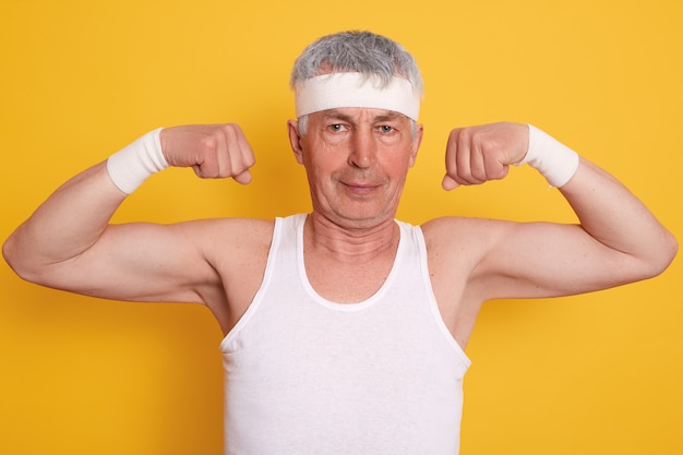 Elderly man dresses white headband showing his biceps and power,, posing against yellow wall after working out