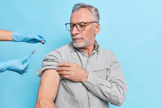 老人はコロナウイルスに対するワクチン接種を受けますワクチンが眼鏡をかけている注射器を注意深く見ます