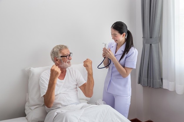 老人の健康的な上昇は、ナーシングホームの寝室で幸せな看護師と一緒に親指を立てる