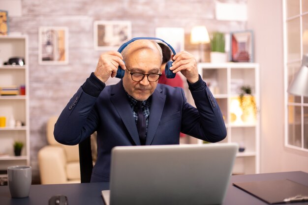 音楽を聴き、現代のラップトップで作業している頭にヘッドフォンを持っている60代の老人