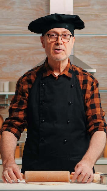 Elderly man rolling dough at home infront of video camera explaining recipe step by step. Retired blogger chef influencer using internet technology communicating on social media with digital equipment