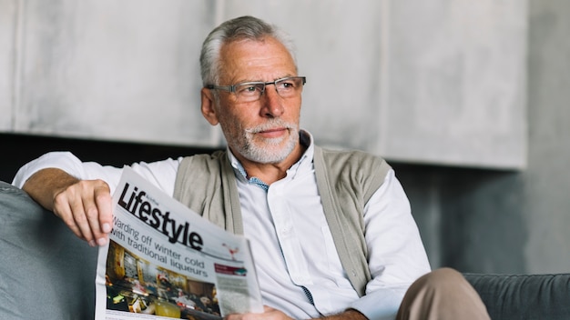 An elderly man sitting on sofa holding newspaper in his hand looking away