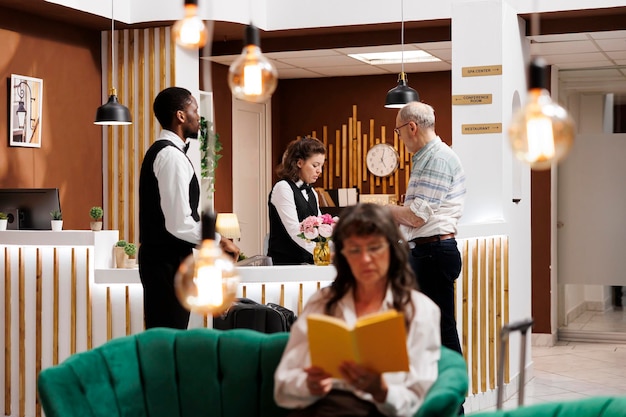 Free photo elderly man visitor at the front desk of the hotel lobby, greeted by pleasant multiethnic employees. while waiting for check-in, a senior woman sits on plush sofa and reads a book in cozy lounge area.