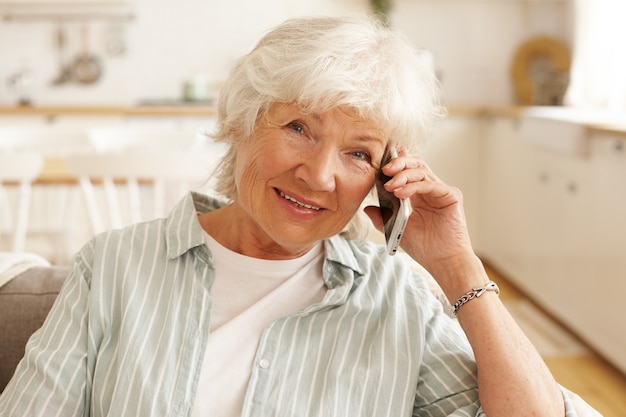Пожилая зрелая европейская женщина в полосатой рубашке разговаривает по телефону через онлайн-приложение, используя бесплатное беспроводное высокоскоростное подключение к Интернету дома, смотрит с веселой улыбкой