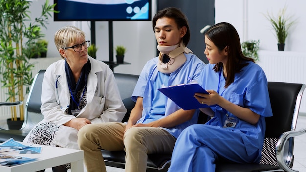 Free photo elderly medic explaining medical expertise with assistant in hospital waiting room, discussing health care treatment with injured patient. asian man wearing cervical neck collar during consultation