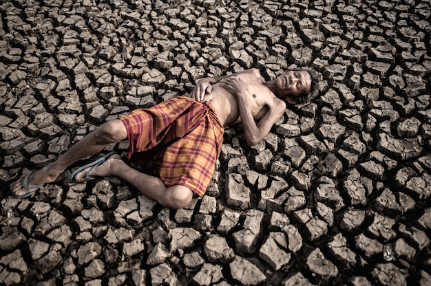 Elderly men lay flat, hands placed on the belly on dry and cracked soil, global warming