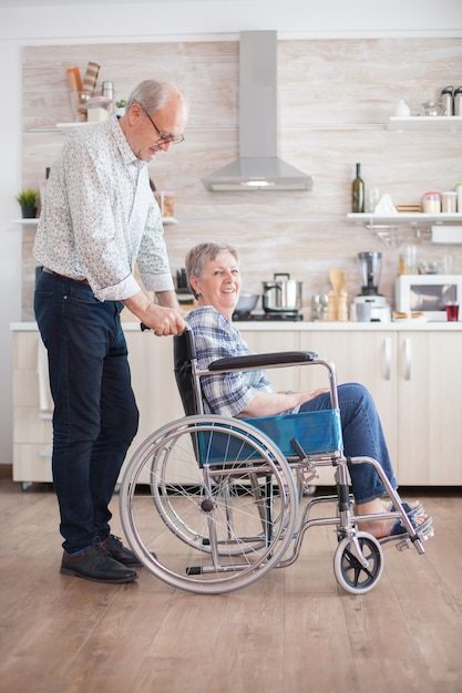 elderly retired man helping his wife with walking disability. Disabled senior woman sitting in wheelchair in kitchen looking through window. Living with handicapped person. Husband helping wife with d