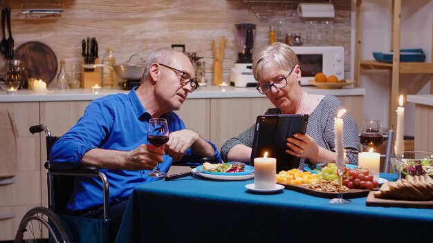 Elderly retired woman showing tablet to paralyzed man, scrolling and showing photos. Imobilized handicapped senior husband browsing on phone enjoying the festive male, drinking a glass of red wine.