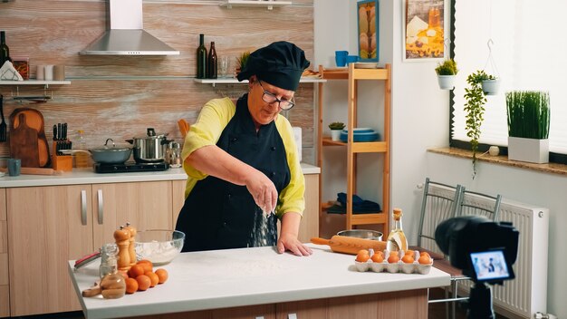 Elderly social media influencer cooking bread using camera on tripod sitting in the kitchen. Retired blogger chef using internet technology communicating, shooting blogging with digital equipment