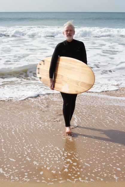 Free photo elderly surfer holding big surfboard. cheerful caucasian man in wetsuit enjoying rest actively spending time when surfing in sea. sport activity and vacation of aged people concept