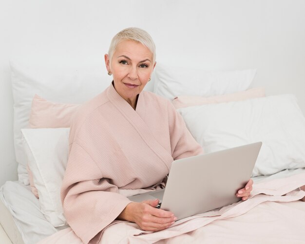 Пожилая женщина в халате позирует на кровати, держа ноутбук