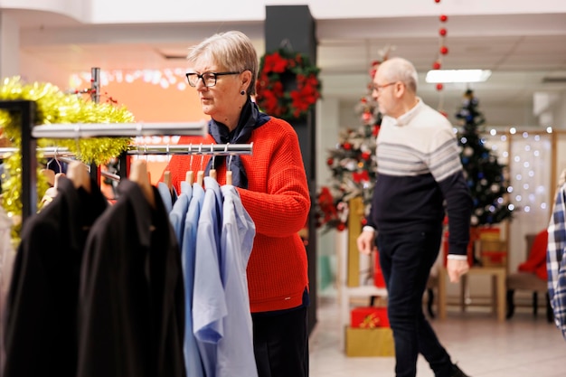 Foto gratuita una donna anziana che compra regali per la cena natalizia, alla ricerca di regali nel centro commerciale durante la stagione delle vendite. una persona anziana che cerca vestiti sugli scaffali, controlla i tessuti.