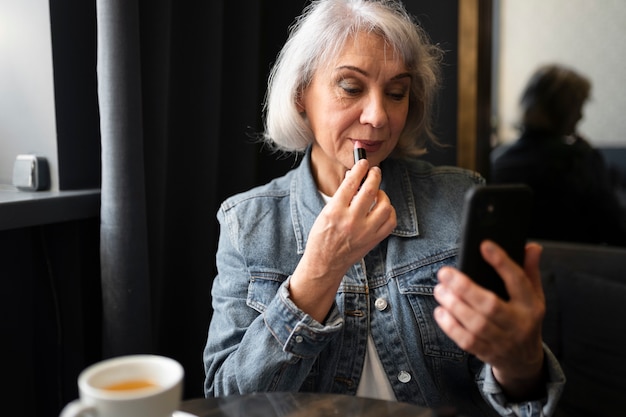 Free photo elderly woman drinking coffee at a cafe and refreshing her make up using lipstick