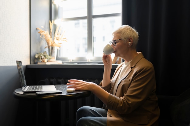 Elderly woman drinking coffee at a cafe while looking at her laptop