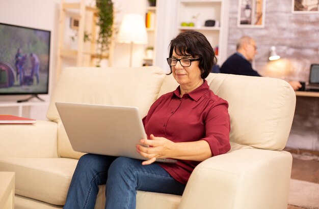 노트북을 사용하여 소파에 앉아 있는 60대 할머니, 신기술을 사용하는 노인