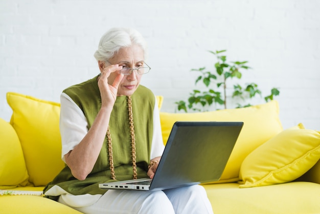 An elderly woman sitting on yellow sofa looking at laptop