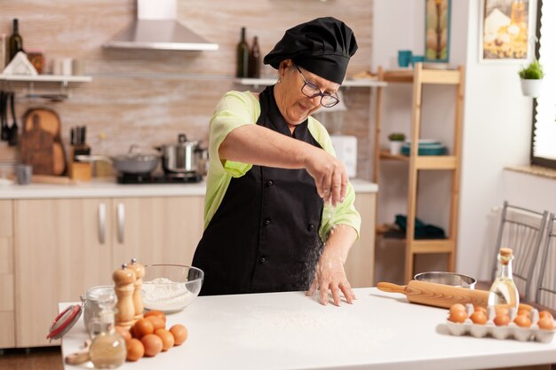 小麦粉を使用して家庭の台所のテーブルに小麦粉を広げておいしいクッキーを作る年配の女性