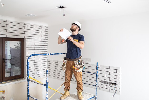 Electrician Builder with beard worker in a white helmet at work, installation of lamps at height. Professional in overalls with a drill on the repair site.