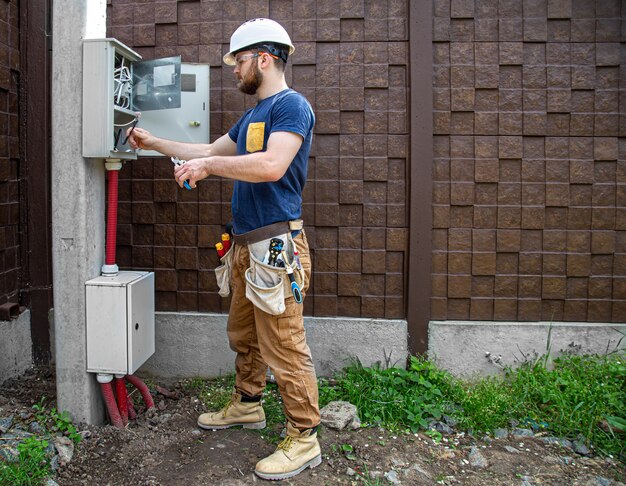 仕事中の電気技師ビルダーは、産業用配電盤の胴体の電線のケーブル接続を調べます。電気技師のツールを使用したオーバーオールのプロ。