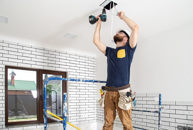 Electrician Builder at work, installation of lamps at height. Professional in overalls with a drill on the repair site.