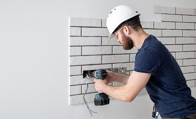 An electrician construction worker in overalls with a drill during the installation of sockets. Home renovation concept.