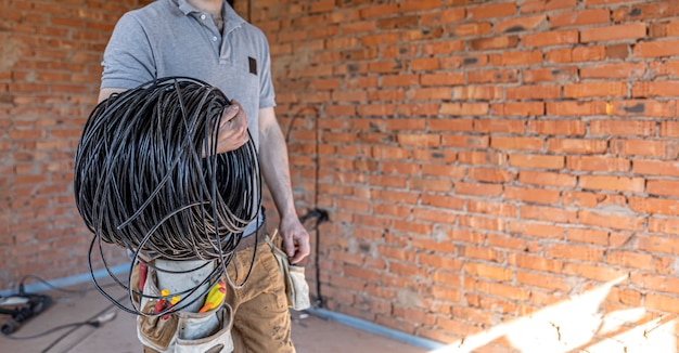 An electrician in a hard hat looks at the wall while holding an electric cable