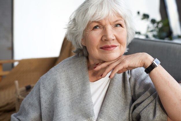 Elegance, age, beauty and people concept. Indoor image of charming elegant senior mature woman enjoying free time at home, sitting on couch in stylish living room interior, smiling happily