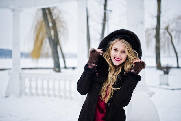 冬の雪の日にポーズをとった毛皮のコートと赤いイブニングドレスの優雅なブロンドの女の子