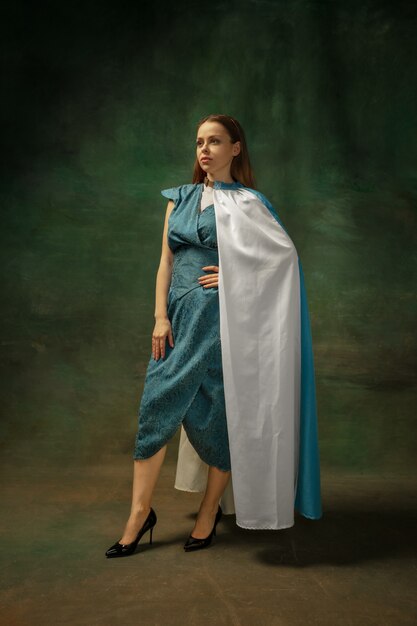 Elegance posing. Portrait of medieval young woman in blue vintage clothing on dark background. Female model as a duchess, royal person. Concept of comparison of eras, modern, fashion, beauty.