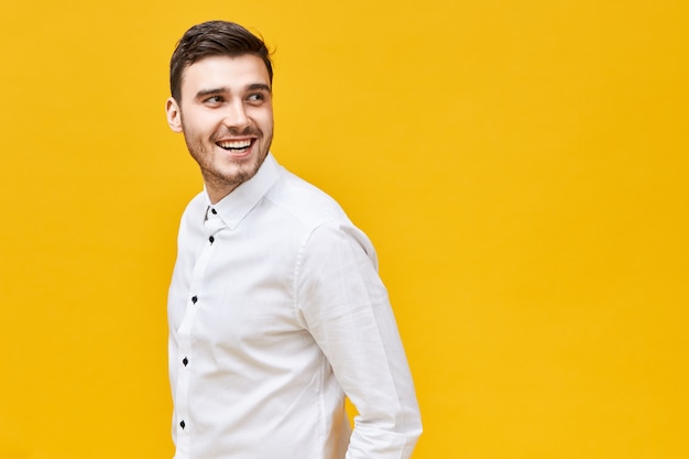 Elegance, style and masculinity concept. Attractive trendy looking young brunette man with bristle and happy smile expressing confidence posing isolated at yellow  wall, turning head round