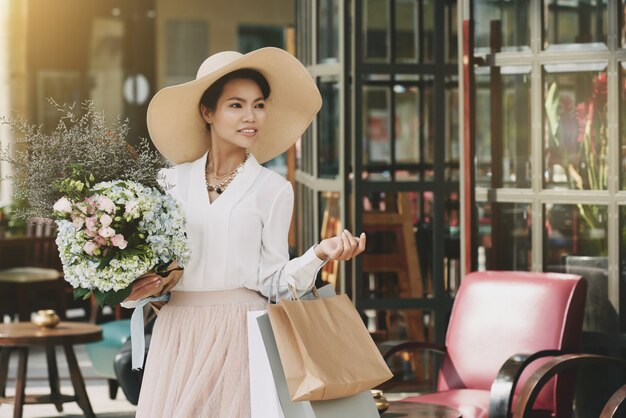 Изящная азиатская леди, выходящая из кафе с хозяйственными сумками и цветочным букетом