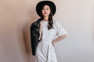 Элегантная азиатская женщина в белом платье смотря камеру. уверенно китаянка в шляпе, держащей кожаную куртку.