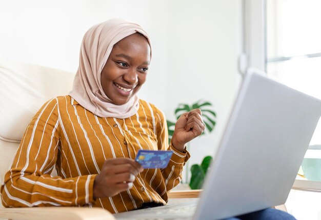 모바일 노트북을 사용하여 집 거실에서 온라인 쇼핑 정보를 검색하는 우아한 매력적인 이슬람 여성 온라인 쇼핑을 통해 제품을 구매하는 행복한 여성의 초상화 신용 카드로 지불