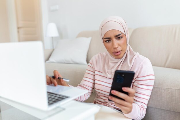 휴대폰과 노트북을 사용하여 집 거실에서 온라인 쇼핑 정보를 검색하는 우아한 매력적인 이슬람 여성 온라인 쇼핑을 통해 제품을 구매하는 행복한 여성의 초상화