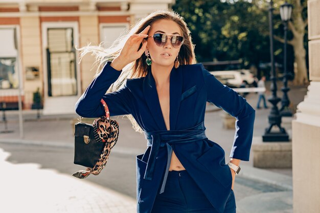 Elegant attractive woman wearing blue stylish suit and sunglasses walking in street holding handbag