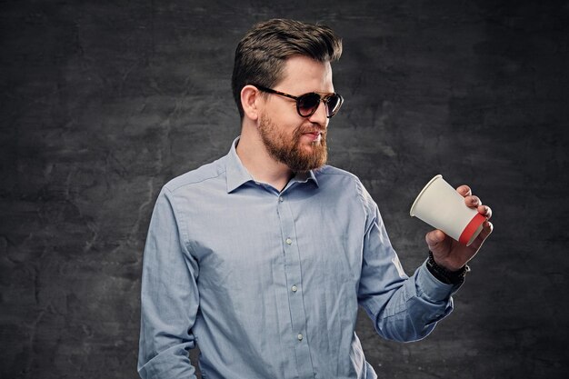 エレガントなひげを生やしたヒップスターの男性は、紙のコーヒーカップを保持しています。