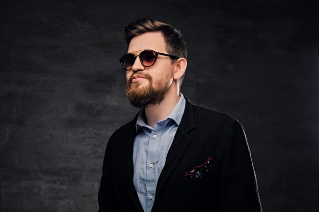 An elegant bearded male dressed in cotton velvet suit and sunglasses over grey background.