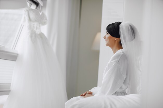 A elegant and beautiful bride at home sitting in a bed near window 