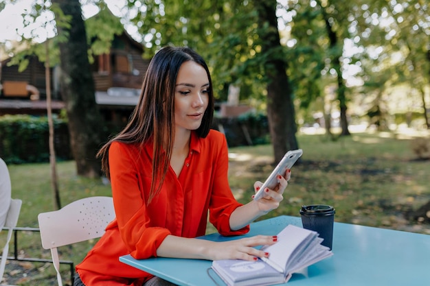 Elegant beautiful dark haired woman in red stylish blouse is smiling widely looks away and sits by little table in street cafe Woman is sitting outdoor with phone in the park