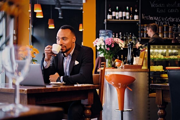 An elegant black American male dressed in a suit drink coffee in a restaurant and working with laptop.