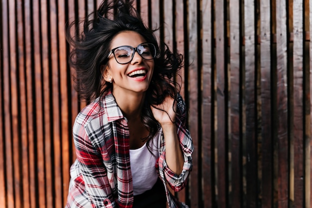 Elegant black-haired girl dancing on wooden wall. Charming latin woman in glasses expressing postivie emotions and laughing.