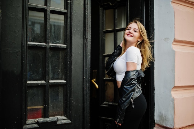 Elegant blonde girl wear on black leather jacket posing at streets of town background old doors