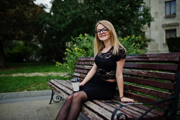 Elegant blonde girl wear on black posing at streets of town sitting on bench with cup of coffee at hands