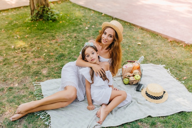 Elegante donna bionda si trova sul manto bianco con cesto di mele nel fine settimana. ritratto all'aperto della ragazza allegra e sua madre che godono del bel tempo nel parco.