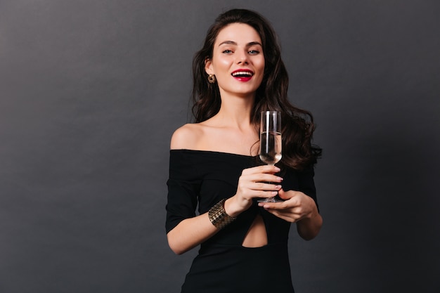 Элегантная голубоглазая девушка с красной помадой улыбается и держит бокал шампанского на черном фоне.
