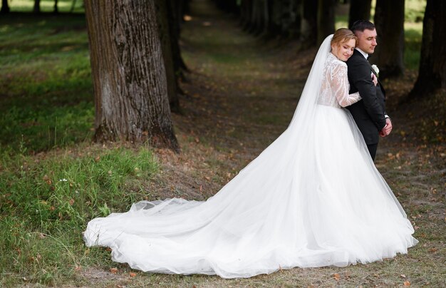 Элегантная невеста в свадебном платье, стоящая позади красивого жениха в саду