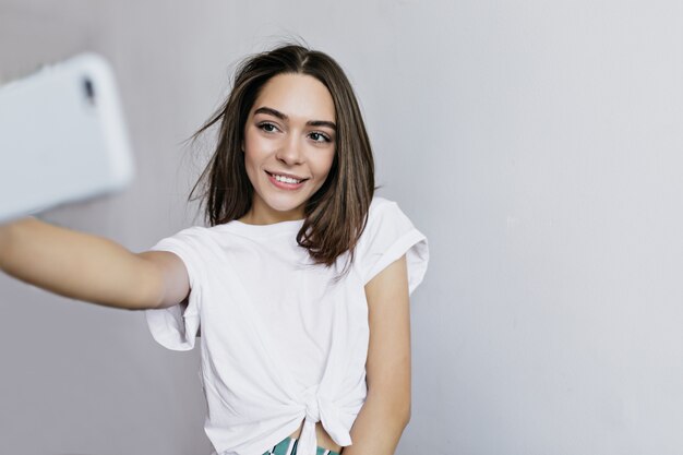 Elegant brown-haired girl holding cell and taking picture of herself. Interested tanned young woman using smartphone for selfie.