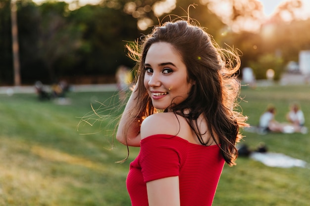 夏の日に公園でポーズをとるエレガントなブルネットの少女。自然の景色を楽しむ赤いドレスを着た陽気な若い女性。